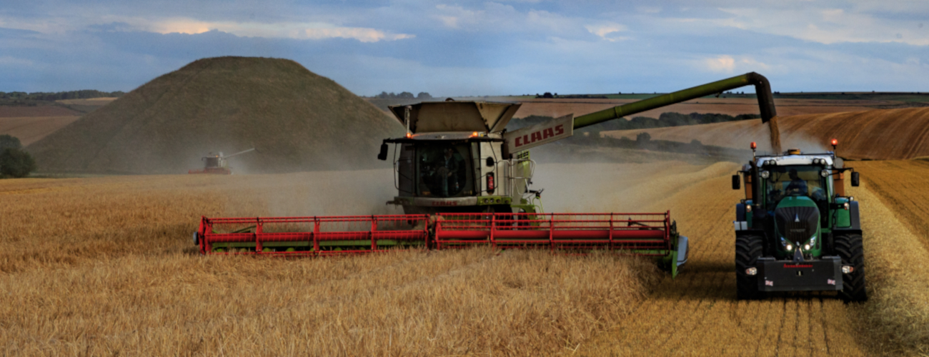 foreground: a combine harvester emptying grain into a trailer, Background: silbury hill